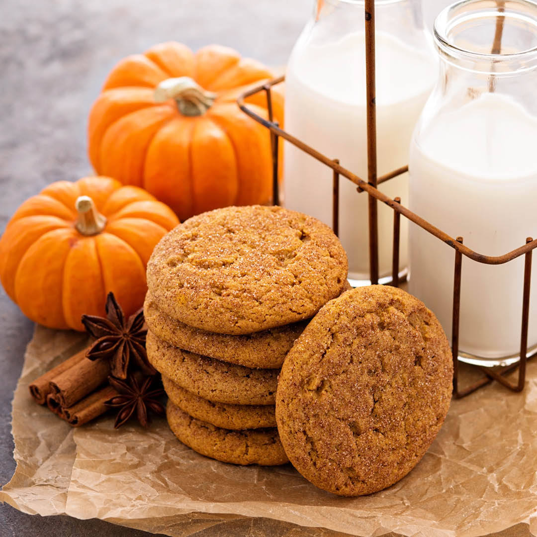 A stack of pumpkin cookies on parchment paper next to two small pumpkins, cinnamon sticks, star anise, and two glass bottles of milk in a metal holder.