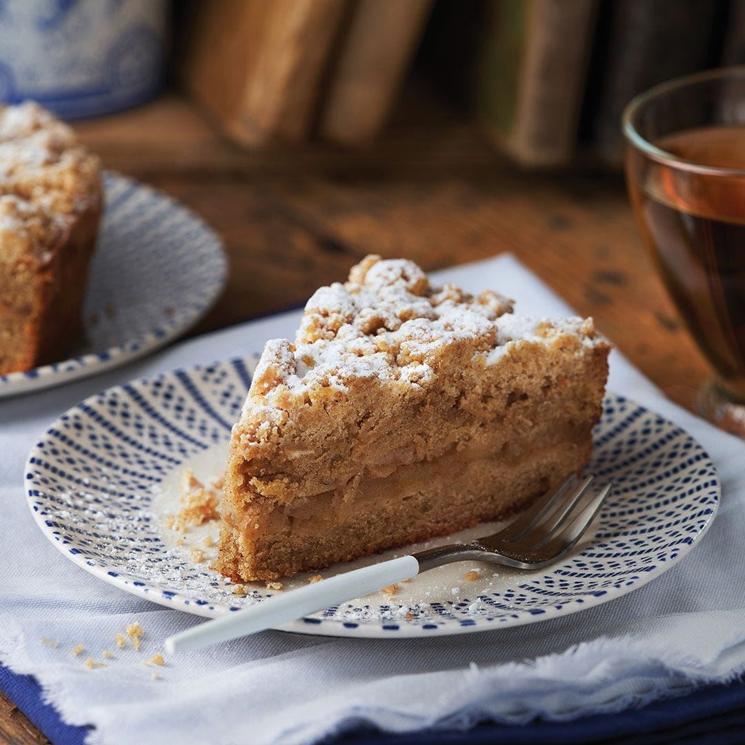 A slice of crumb-topped apple cake dusted with powdered sugar sits on a patterned plate with a fork. A glass of tea and another plate with cake are in the background.