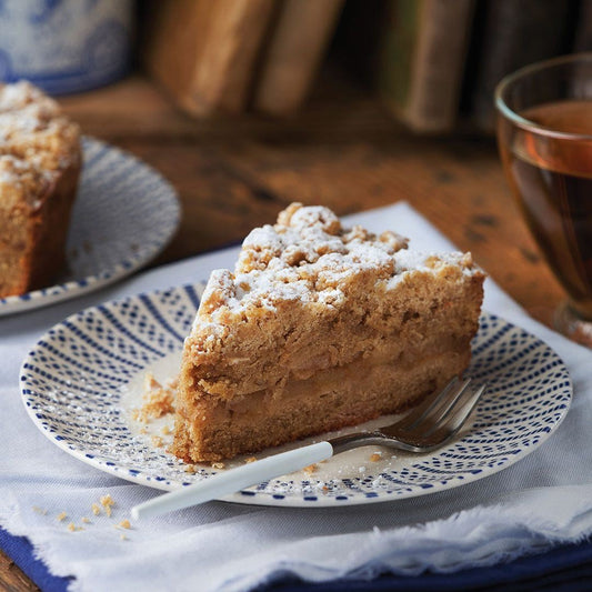 A slice of crumb-topped apple cake dusted with powdered sugar sits on a patterned plate with a fork. A glass of tea and another plate with cake are in the background.
