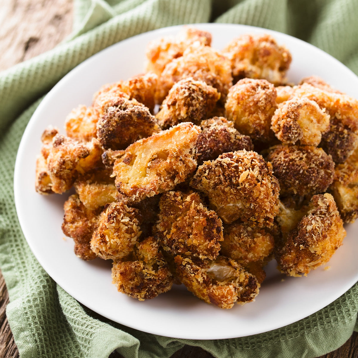 A white plate piled with crispy, golden-brown breaded cauliflower florets sits on a green cloth napkin. The cauliflower appears freshly baked or fried, with a crunchy coated texture.