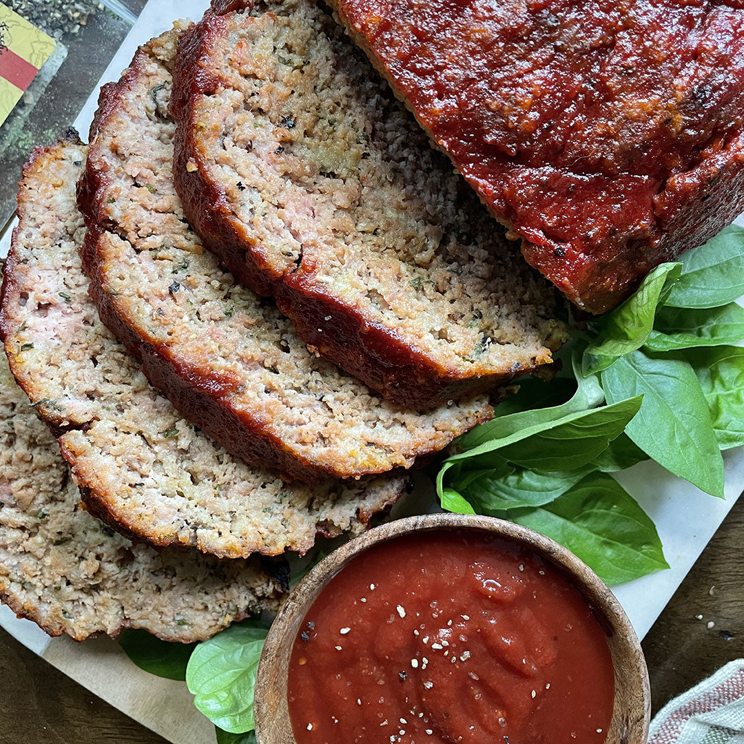 Sliced meatloaf topped with tomato sauce is arranged on leafy greens, with a small wooden bowl of seasoned tomato sauce on the side.