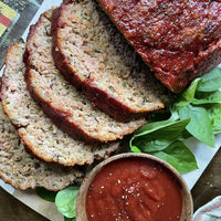 Sliced meatloaf topped with tomato sauce is arranged on leafy greens, with a small wooden bowl of seasoned tomato sauce on the side.