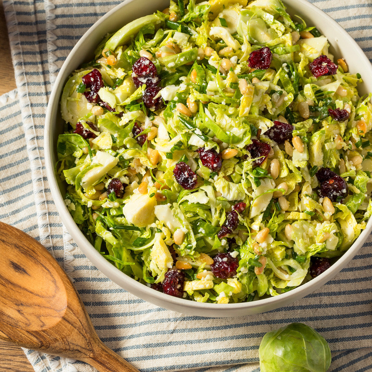 A bowl of shredded Brussels sprout salad with dried cranberries, pine nuts, and parmesan cheese, placed on a striped cloth next to a wooden serving spoon.