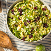 A bowl of shredded Brussels sprout salad with dried cranberries, pine nuts, and parmesan cheese, placed on a striped cloth next to a wooden serving spoon.