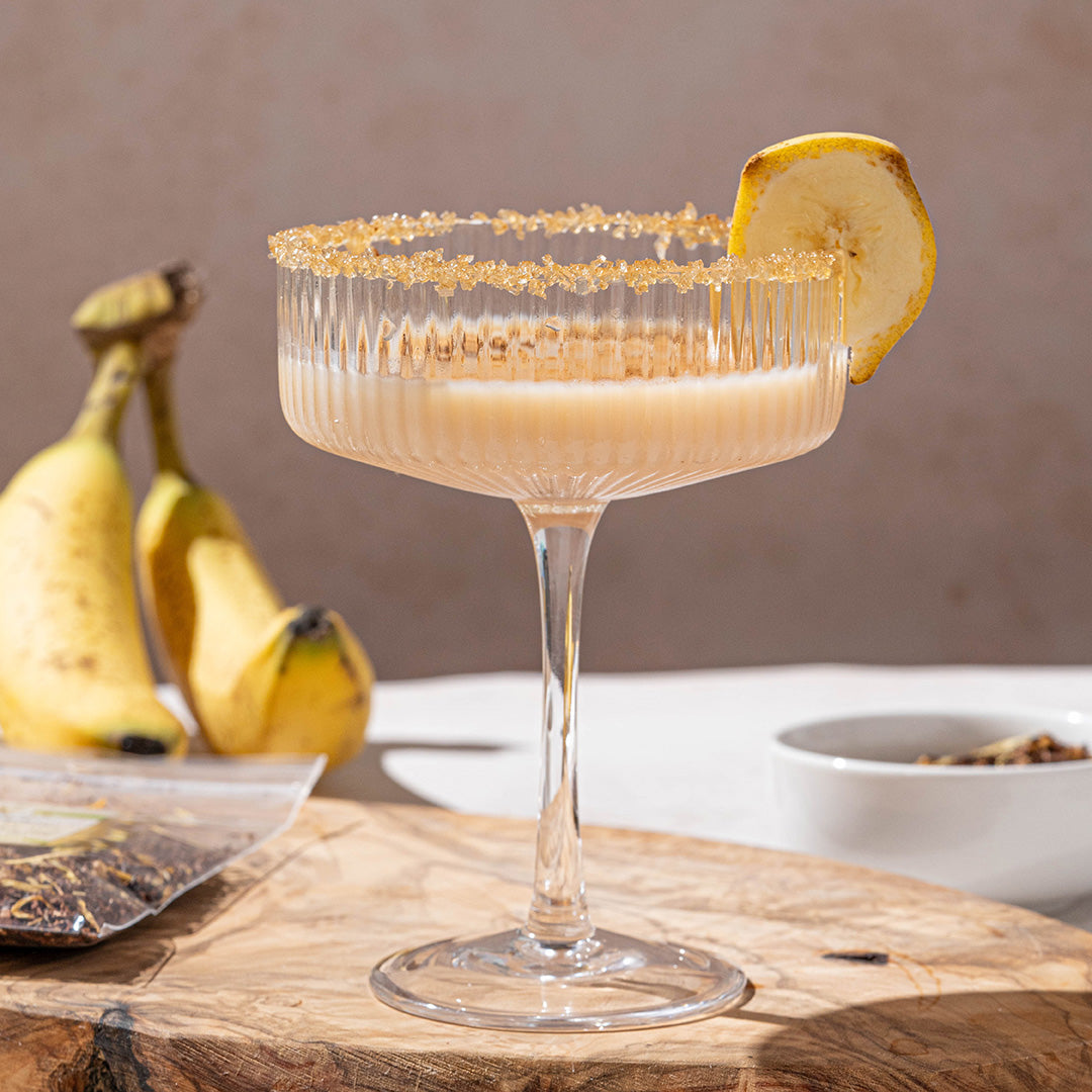 A cocktail glass with a sugared rim and a banana slice garnish, filled with a creamy drink, sits on a wooden board. In the background, bananas and a white bowl are visible.
