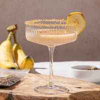 A cocktail glass with a sugared rim and a banana slice garnish, filled with a creamy drink, sits on a wooden board. In the background, bananas and a white bowl are visible.