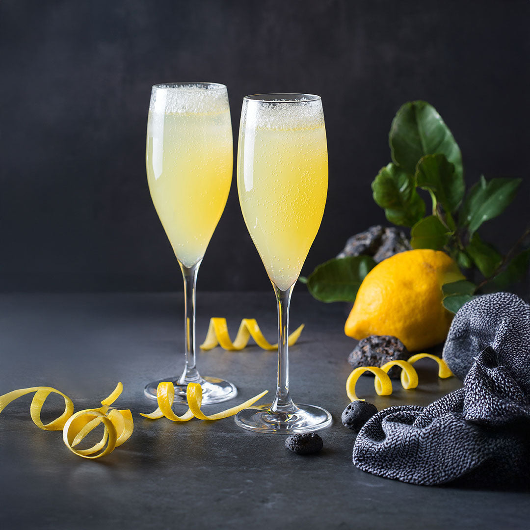 Two champagne flutes filled with a yellow, fizzy cocktail sit on a dark surface, surrounded by lemon peels, a whole lemon with leaves, decorative stones, and a textured dark cloth.