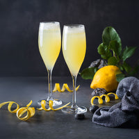 Two champagne flutes filled with a yellow, fizzy cocktail sit on a dark surface, surrounded by lemon peels, a whole lemon with leaves, decorative stones, and a textured dark cloth.