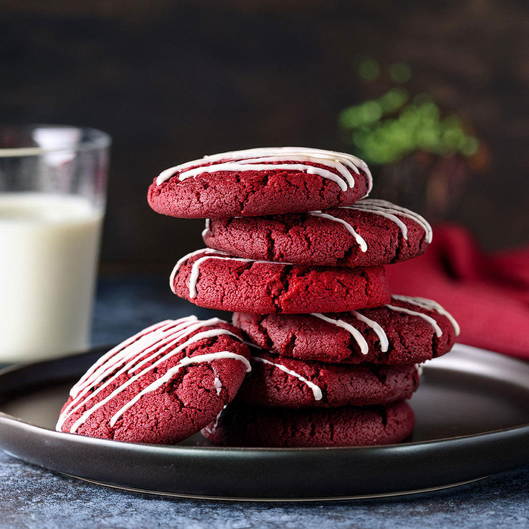 A stack of red velvet cookies with white icing drizzle on a dark plate, with a glass of milk in the background and a blurred plant visible.