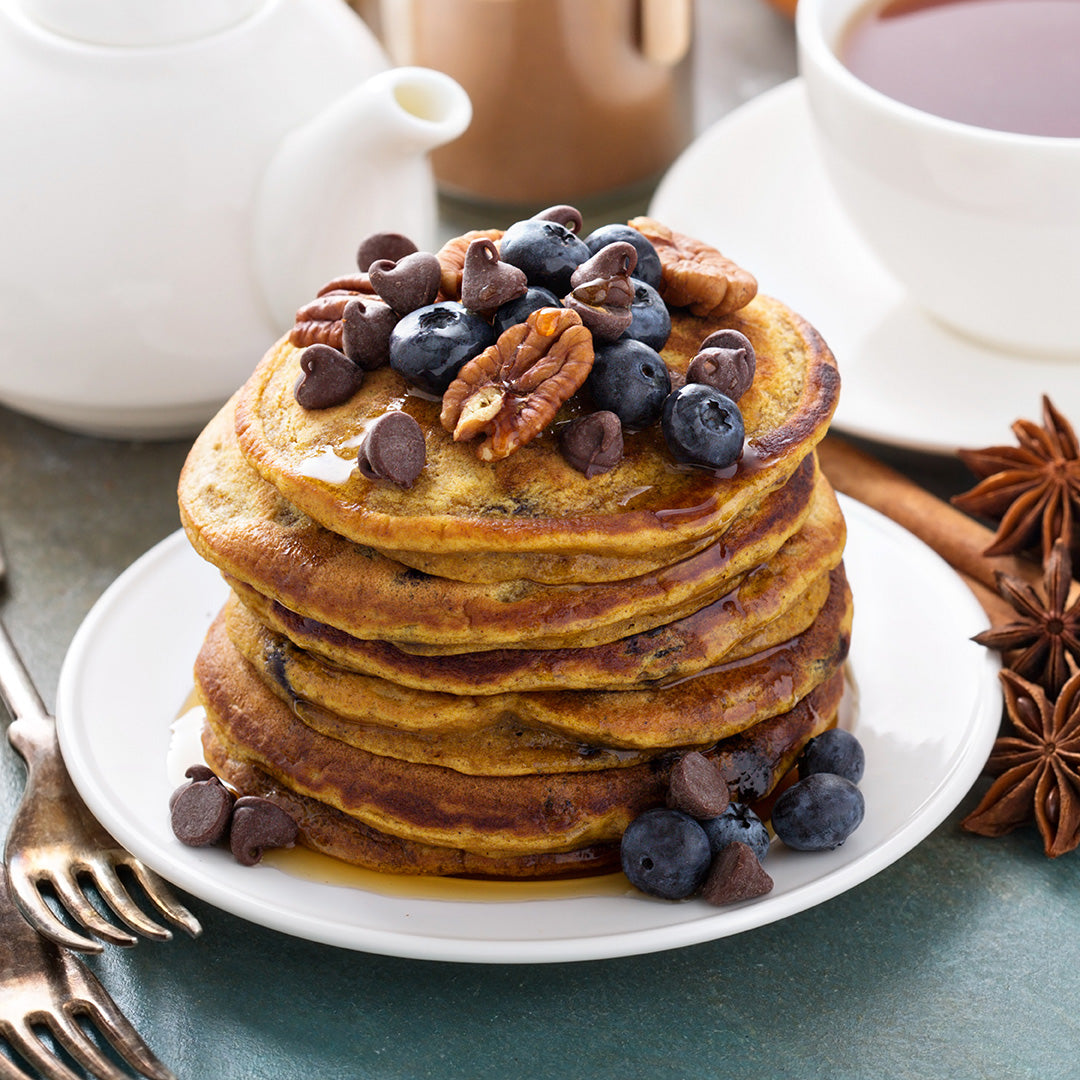 A stack of pancakes topped with blueberries, chocolate chips, and pecans sits on a white plate beside a cup of tea and a white teapot, with a fork nearby on the table.