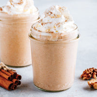 Two glass jars filled with a creamy, light brown smoothie, topped with whipped cream and sprinkled with cinnamon. Cinnamon sticks and walnuts are placed nearby on a light surface.