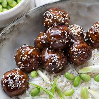 A close-up of glazed meatballs topped with sesame seeds, served on white rice with green edamame and sliced scallions in a gray bowl. A dish of more edamame is visible in the background.