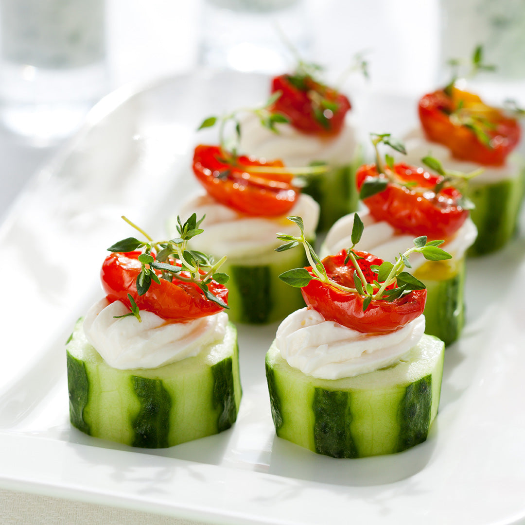 Cucumber slices topped with a swirl of creamy cheese, roasted cherry tomatoes, and fresh thyme are arranged neatly on a white plate.