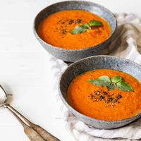 Two bowls of Tuscan Tomato Soup (Kit), topped with fresh basil and cracked black pepper, sit on a striped napkin with two spoons nearby, inviting you to enjoy the classic flavors of Tuscany.