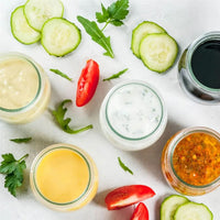 Five jars of different salad dressings are arranged on a white surface, surrounded by cucumber slices, tomato wedges, arugula, and parsley for a fresh and colorful presentation.