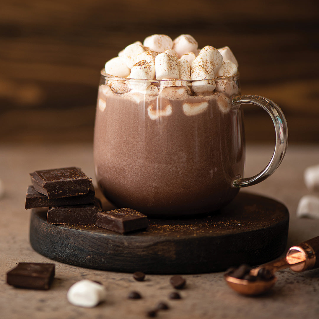 A glass mug of hot chocolate topped with mini marshmallows and a sprinkle of cocoa powder sits on a wooden coaster. Chocolate pieces, coffee beans, and marshmallows are scattered around on the table.