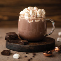 A glass mug of hot chocolate topped with mini marshmallows and a sprinkle of cocoa powder sits on a wooden coaster. Chocolate pieces, coffee beans, and marshmallows are scattered around on the table.