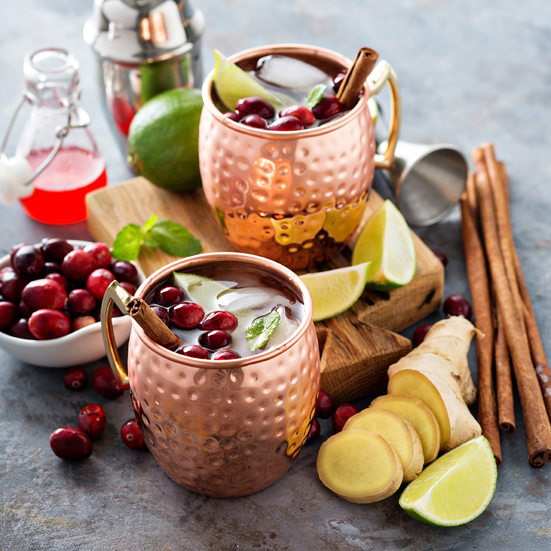 Two copper mugs filled with a drink garnished with cranberries, lime wedges, mint, and cinnamon sticks. Surrounding the mugs are fresh ginger slices, whole cranberries, lime, cinnamon sticks, and a cocktail shaker.