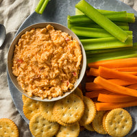 A plate with a bowl of pimento cheese dip, surrounded by celery sticks, carrot sticks, and round butter crackers. The platter is set on a gray surface with a beige napkin.