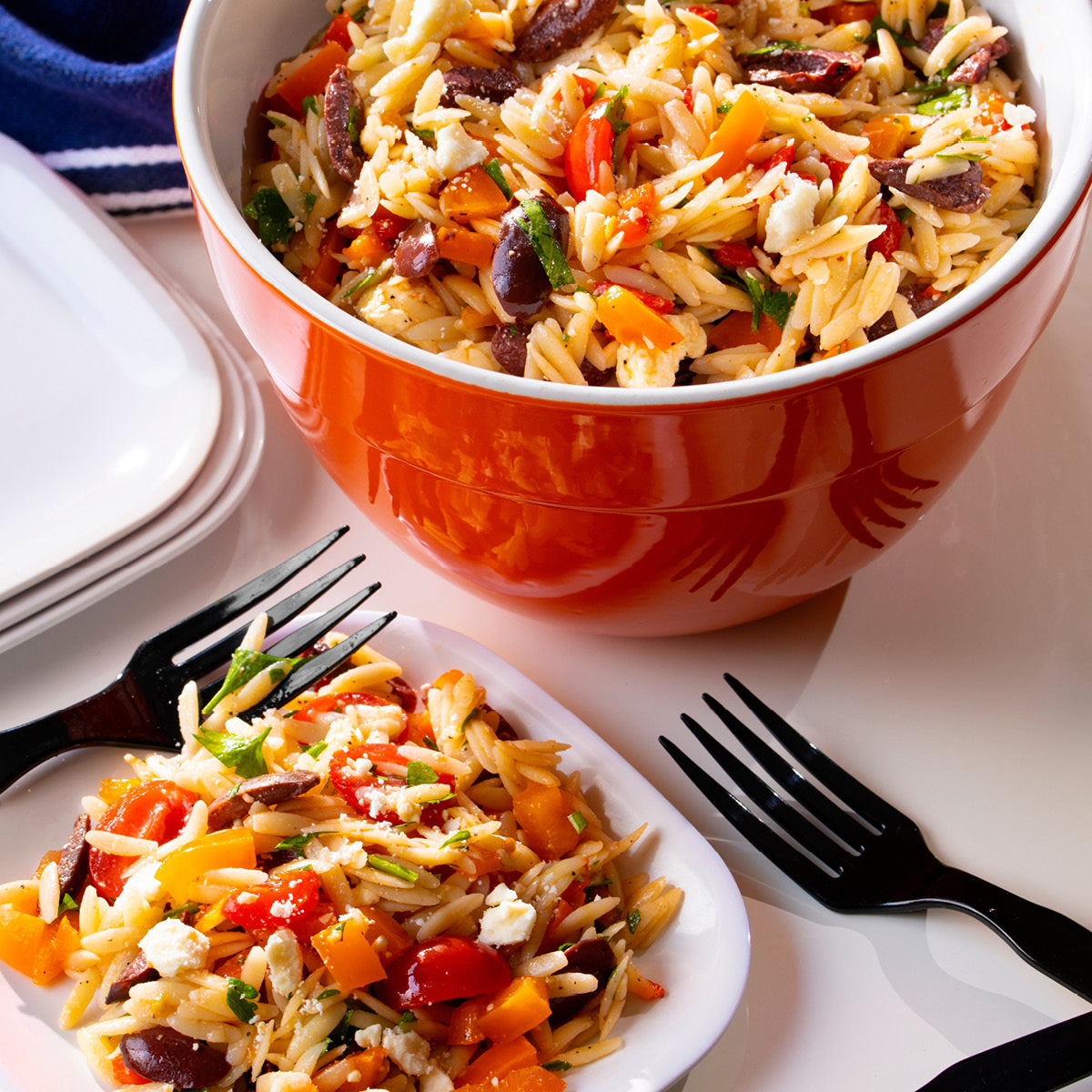 A large orange bowl and a small plate filled with orzo salad mixed with chopped bell peppers, olives, feta cheese, and herbs. Two black forks lie nearby on a white table.