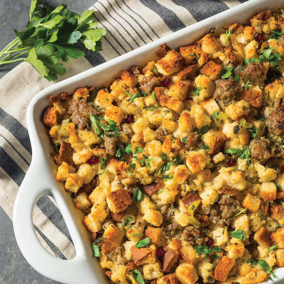 A white casserole dish filled with golden-brown stuffing made of bread cubes, sausage, herbs, and garnished with fresh parsley, resting on a striped cloth with parsley leaves nearby.
