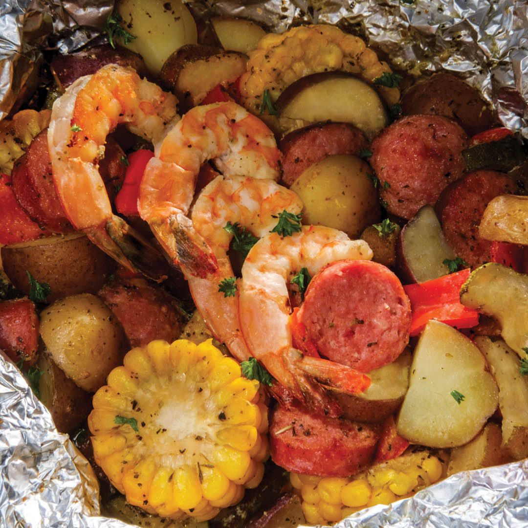 Shrimp, sliced sausage, corn on the cob, potatoes, and bell peppers cooked together in foil, garnished with herbs.