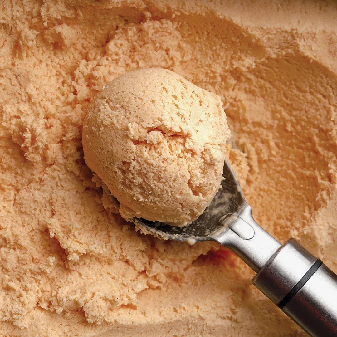 Close-up of a metal ice cream scooper lifting a scoop of creamy, light orange ice cream from a larger container or tray of the same ice cream.