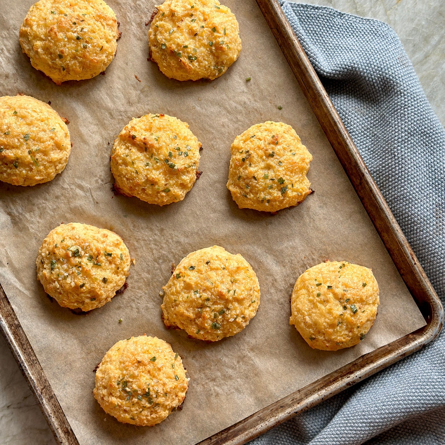 A baking tray lined with parchment paper holds ten golden, round cheese biscuits, sprinkled with herbs. The tray rests on a gray textured cloth on a light countertop.