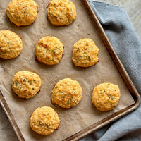 A baking tray lined with parchment paper holds ten golden, round cheese biscuits, sprinkled with herbs. The tray rests on a gray textured cloth on a light countertop.