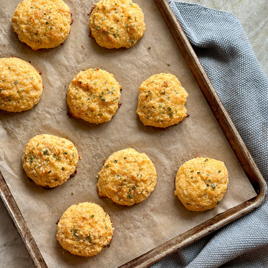 A baking tray lined with parchment paper holds ten golden, round cheese biscuits, sprinkled with herbs. The tray rests on a gray textured cloth on a light countertop.
