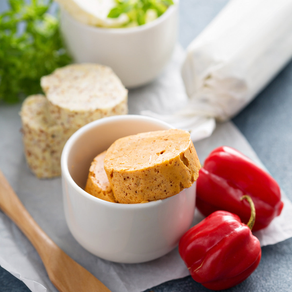A small white bowl with two slices of seasoned compound butter sits on parchment paper next to red bell peppers. More butter, herbs, and a wooden spoon are in the background.