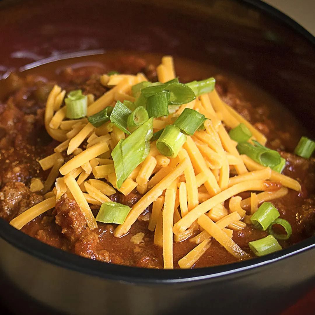 A bowl of chili topped with shredded cheddar cheese and chopped green onions. The chili appears thick and hearty, served in a dark-colored bowl.