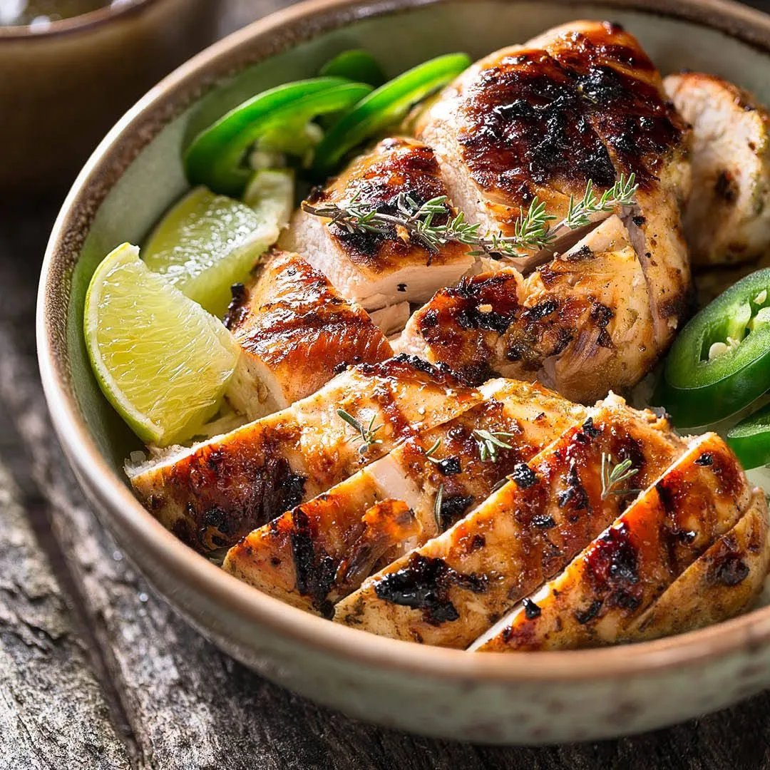 A bowl filled with sliced, grilled chicken breast, garnished with fresh herbs, lime wedges, and sliced jalapeños, placed on a rustic wooden surface.