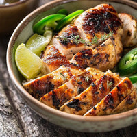 A bowl filled with sliced, grilled chicken breast, garnished with fresh herbs, lime wedges, and sliced jalapeños, placed on a rustic wooden surface.