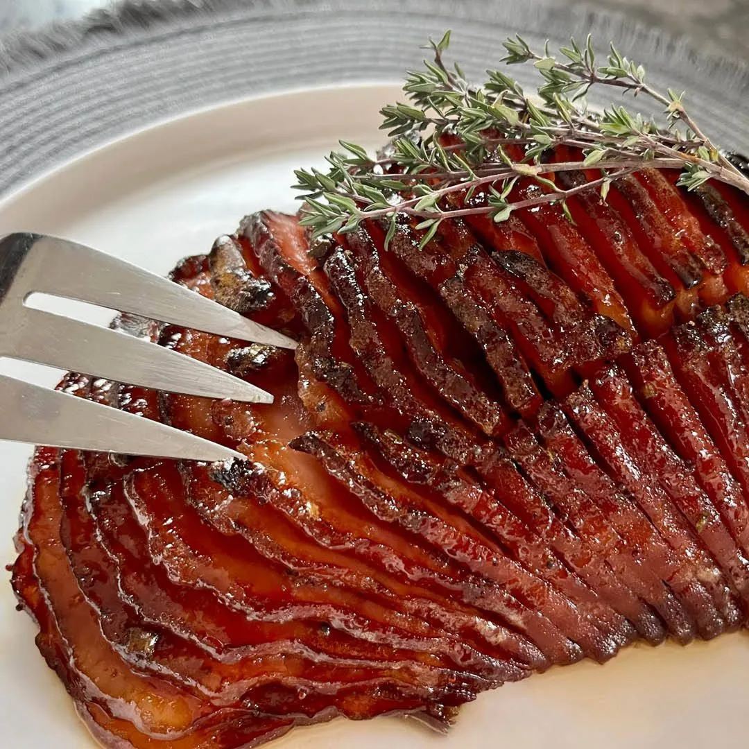A close-up of a glazed, spiral-cut ham garnished with sprigs of fresh thyme on a white plate, with a silver meat fork inserted into the ham.