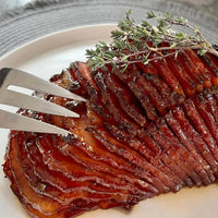 A close-up of a glazed, spiral-cut ham garnished with sprigs of fresh thyme on a white plate, with a silver meat fork inserted into the ham.
