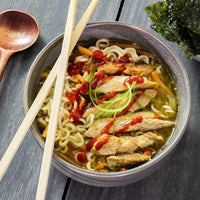 A bowl of ramen with sliced meat, noodles, green garnish, and a drizzle of red sauce, next to wooden chopsticks and a wooden spoon, on a gray wooden table; a dish of seaweed is visible at the side.