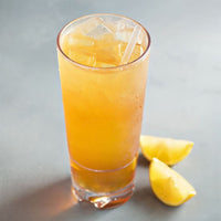 A tall glass filled with an orange-colored iced drink and a clear straw, placed on a gray surface with two lemon wedges beside the glass.