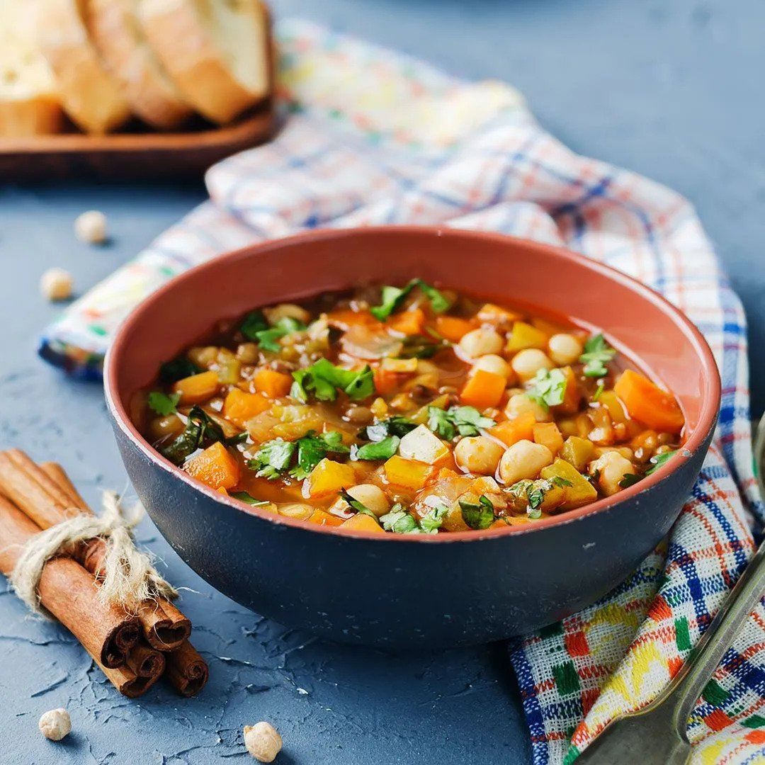 A bowl of vegetable and chickpea stew garnished with fresh herbs sits on a colorful cloth, with bread slices and cinnamon sticks nearby on a textured blue surface.