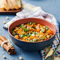 A bowl of vegetable and chickpea stew garnished with fresh herbs sits on a colorful cloth, with bread slices and cinnamon sticks nearby on a textured blue surface.