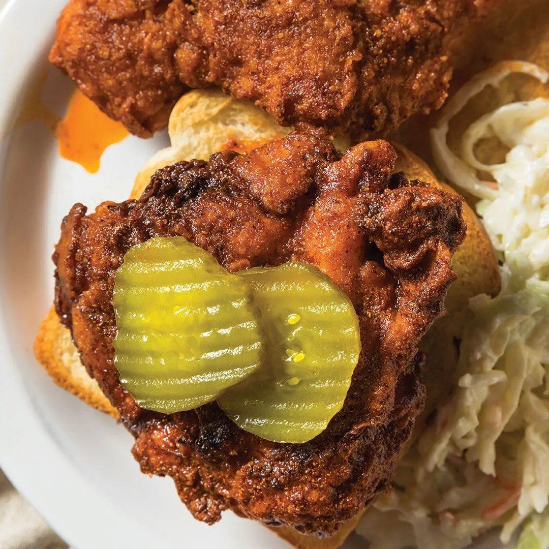A plate with a piece of spicy fried chicken topped with two pickle slices on bread, served next to a side of creamy coleslaw.