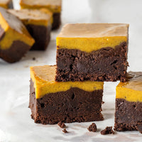 Three rich, fudgy brownies topped with a thick pumpkin layer are stacked on parchment paper, with more brownies blurred in the background. The surface is smooth and glossy, highlighting the dessert’s contrasting layers.