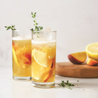 Two tall glasses filled with iced tea, lemon and peach slices, garnished with fresh thyme sprigs. In the background, a wooden board holds a halved lemon and peach.