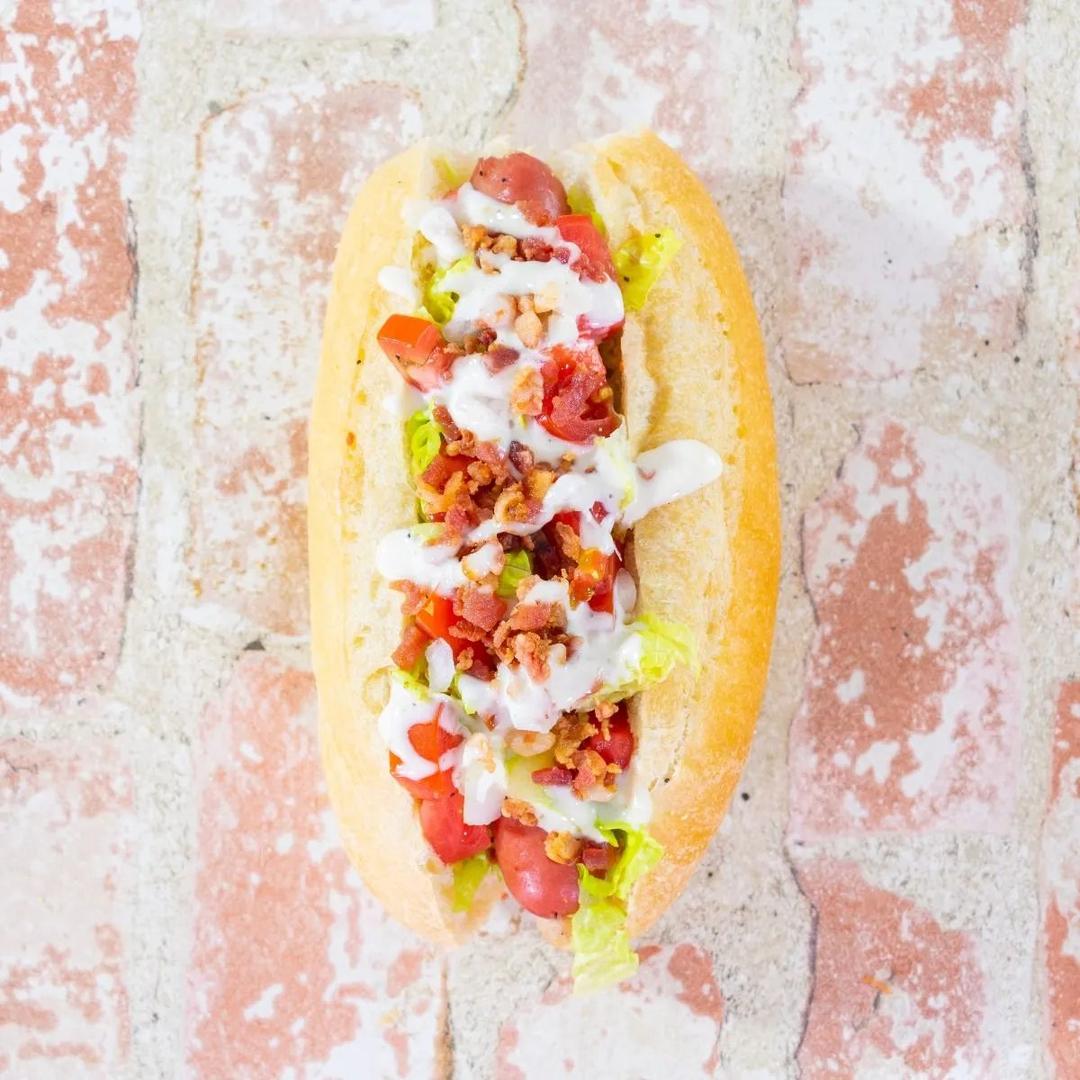 A hot dog in a bun topped with chopped lettuce, tomatoes, bacon bits, and a drizzle of creamy white sauce, placed on a rustic white and red brick background.