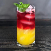 A layered cocktail in a tall glass with yellow liquid at the bottom, red liquid on top, ice cubes throughout, and a mint sprig garnish. The drink is placed on a dark wooden surface.