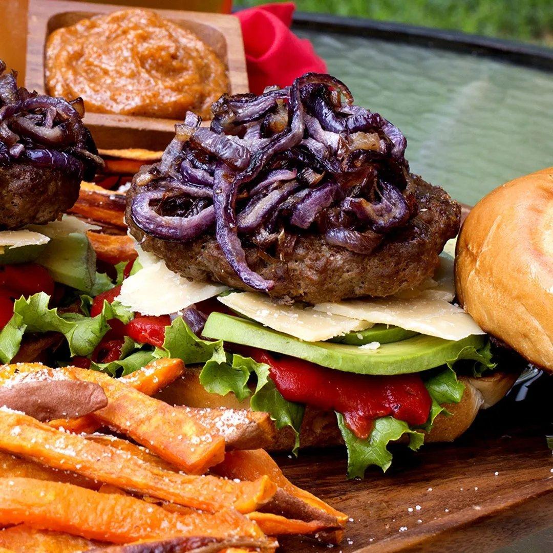 A close-up of a burger topped with grilled onions, avocado slices, lettuce, cheese, and roasted red pepper on a bun, served with sweet potato fries and a small dish of orange dipping sauce.
