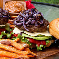 A close-up of a burger topped with grilled onions, avocado slices, lettuce, cheese, and roasted red pepper on a bun, served with sweet potato fries and a small dish of orange dipping sauce.