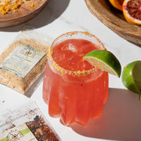 A bright pink cocktail with ice, garnished with a lime wedge and a spiced rim, sits on a white surface surrounded by packets of spices and a wooden plate with blood oranges and lime.