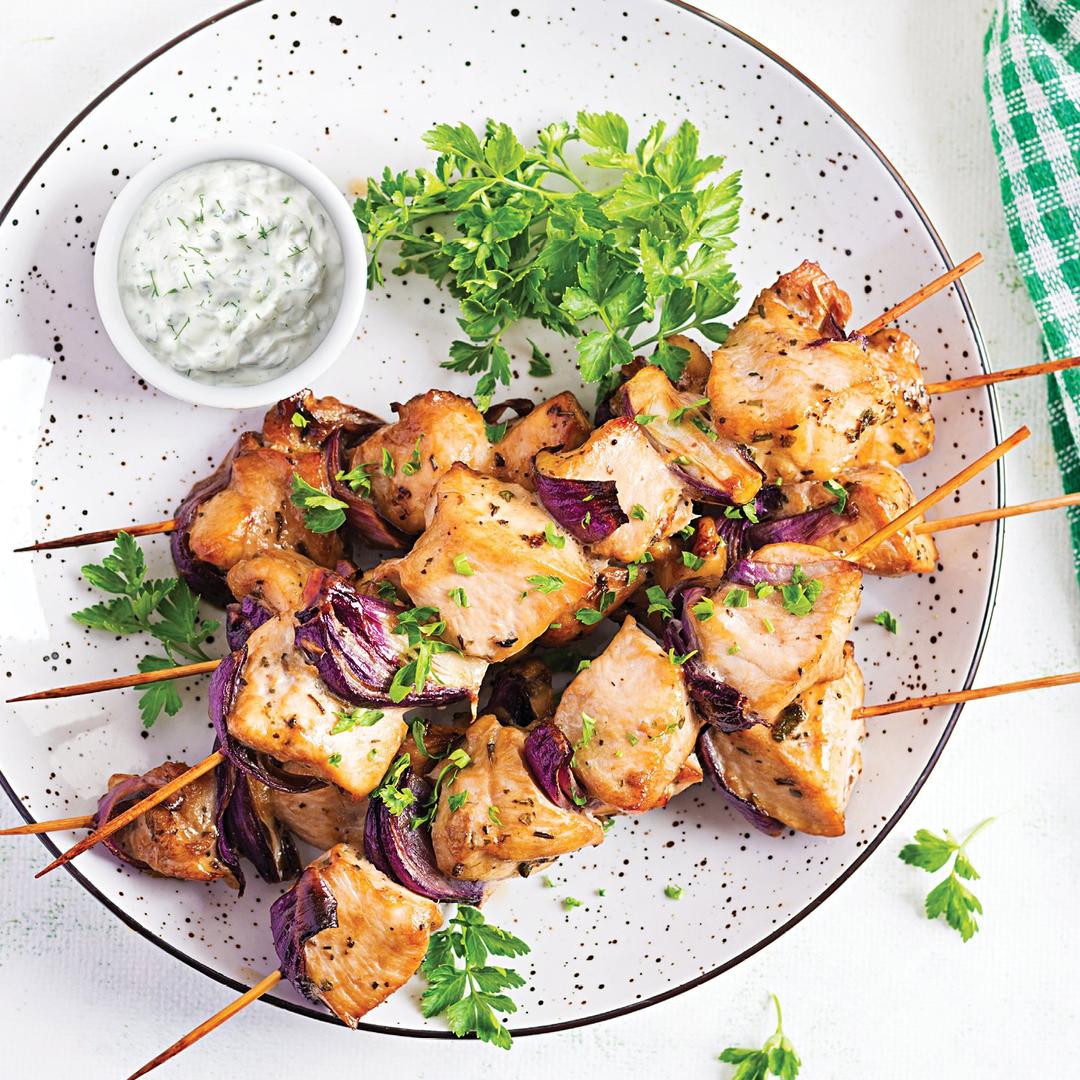 Four grilled chicken and vegetable skewers garnished with chopped herbs are arranged on a white plate. A small bowl of creamy dipping sauce and some fresh parsley are also on the plate.