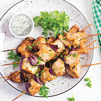 Four grilled chicken and vegetable skewers garnished with chopped herbs are arranged on a white plate. A small bowl of creamy dipping sauce and some fresh parsley are also on the plate.